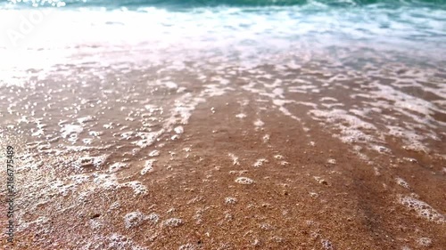 Close-up low-angle shot of ocean water washing over golden sand, leaving shimmering bubbles and white foam under bright daylight.