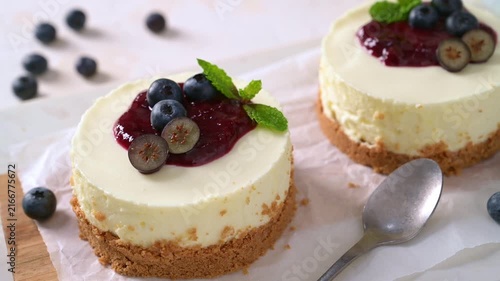Two mini cheesecakes rest on parchment paper, adorned with vibrant berry sauce, fresh blueberries, and mint leaves, illuminated by soft natural lighting.