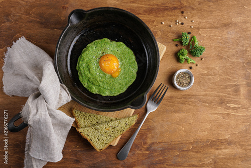 Healthy green fried egg with broccoli in cast iron skillet on wooden table. Top view of a rustic breakfast flat lay with whole grain bread.