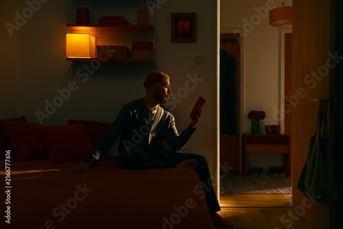 Young man in beanie sitting on bed reading book in cozy dimly lit warm apartment room at evening enjoying peaceful solitary literature study relaxation night hobby inside domestic ambient dark home