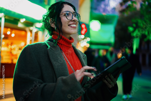 фотографія Happy multiracial female looking up and dreaming while working on tablet in city at night, ambitious student or professional, digital nomad career, authentic ethnic representation