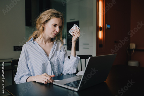 Woman holding a blister pack of pills while using a laptop for an online healthcare consultation at home