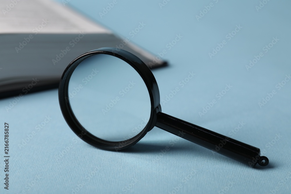 Fototapeta premium Keywords. Magnifying glass and book on light blue background, selective focus. Space for text