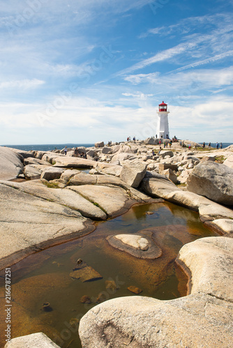 Lighthouse in background and rock formations with water in foreground at Peggy's Cover in summer with blue sky