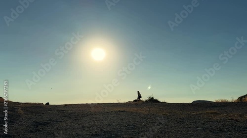 A cinematic video of a woman silhouette walking along a dry barren peninsula during warm summer evening sunset with a vast blue sky and serene coastal atmosphere.