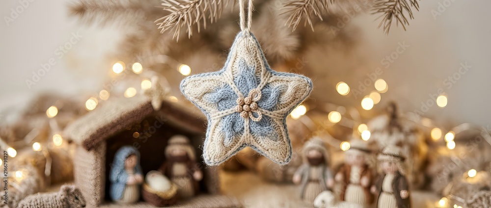Fototapeta premium Knitted star ornament hanging above a nativity scene and glowing Christmas tree lights. Cozy handmade holiday decoration symbolizing faith, family, tradition, and festive celebration.