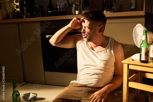 Photography Middle aged Caucasian man sitting on kitchen floor holding head in pain with beer bottles around, representing alcoholism and hangover