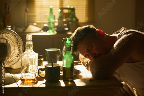 Photography Middle aged Caucasian man sleeping on kitchen table surrounded by empty alcohol bottles, representing substance abuse