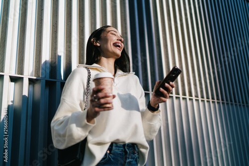 Overjoyed young woman with braces laughing sincerely while holding smartphone and coffee. Girl expressing happiness and success, receiving good news on mobile phone in urban setting, low angle view.