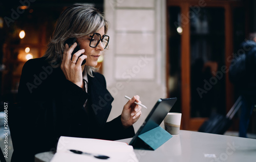 Canvas Print Caucasian businesswoman in stylish glasses talking on mobile phone at terrace table, middle aged female professional using stylus and tablet for work, remote office and digital lifestyle