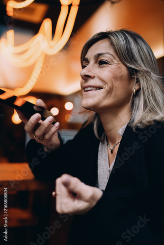 Bild auf Leinwand Smiling mature female using smartphone for voice chat in restaurant, happy woman