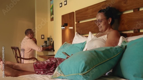 Mature woman using smartphone in bed while man works on laptop