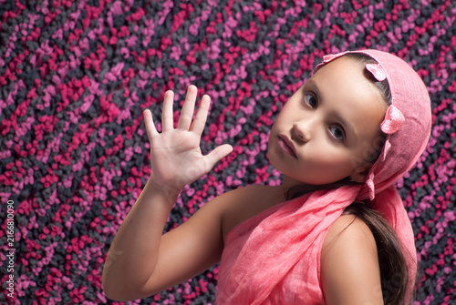 Young girl wearing a pink headscarf and matching dress raising open hand facing camera against a pink textured background