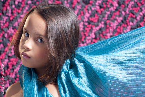 Portrait of a graceful young girl with dark brown hair wrapped in a flowing blue silk scarf against a pink textured background