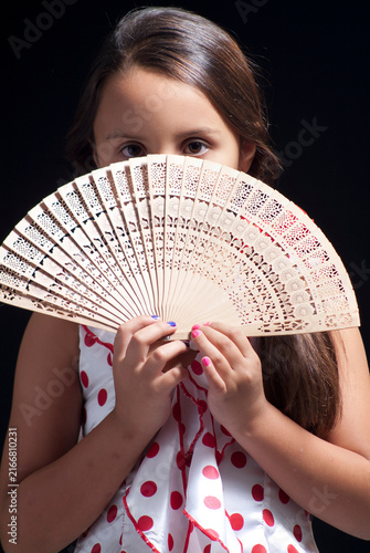 Playful young girl holding a carved wooden folding hand fan covering lower face on black background