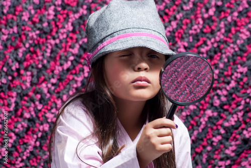 Curious young girl wearing a grey hat looking through a magnifying glass against a pink textured background