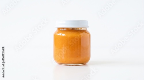 A smooth glass jar containing vibrant orange pureed baby food illuminated under soft bright studio lighting against clean white backdrop.