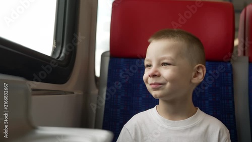 Child seated on train with red headrest blue patterned seat. Natural window light illuminates face. Calm mood, travel theme, warm tones, soft focus foreground