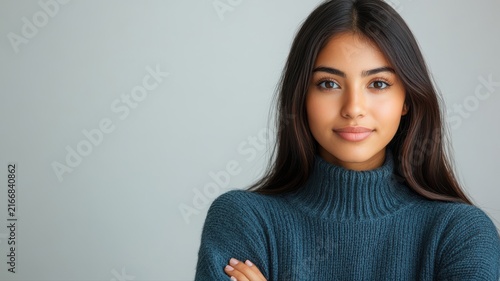 Portrait of a young woman with long dark hair, wearing a cozy blue turtleneck sweater, smiling gently on a plain light gray background