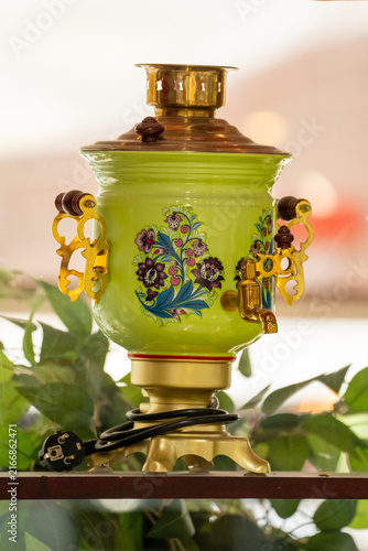 Photography Traditional green metal electric samovar decorated with bright floral patterns and a brass spout, standing on a shelf