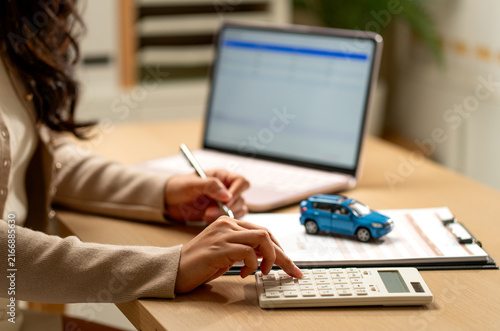 Woman calculating car loan with blue model vehicle on clipboard beside laptop