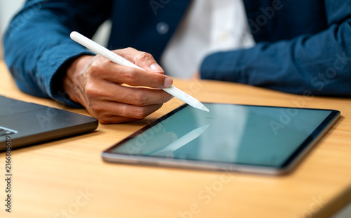 Hand using stylus on tablet with laptop beside on warm wooden desk in natural light