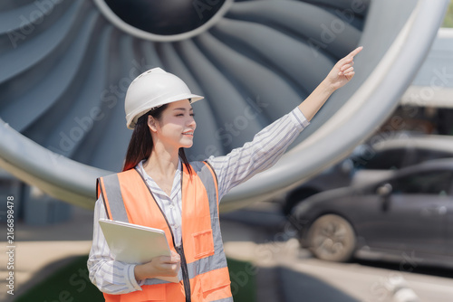 Asian female aeronautical engineer holding laptop and clipboard standing in front of airplane jet engine