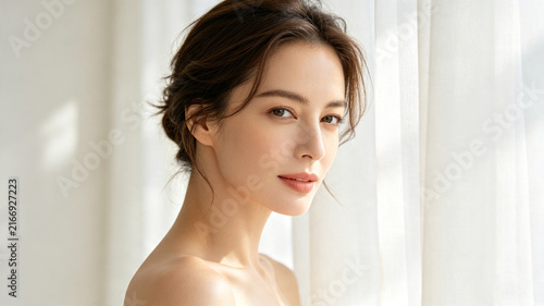 Serene portrait of a beautiful woman with a messy bun and flawless skin, glowing in soft natural light against airy white curtains.