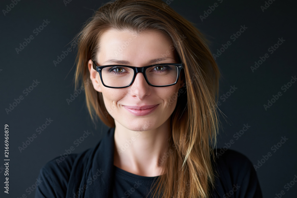 Fototapeta premium Young woman with glasses brown hair brown eyes casual clothing studio portrait friendly smile confident expression smart appearance black background modern style professional headshot