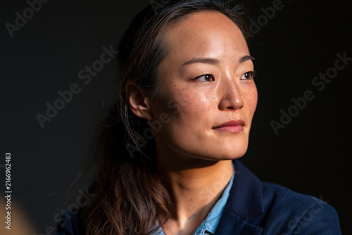 Confident woman portrait Asian woman natural light side view serious expression freckles dark background blue blazer soft shadow contemplation