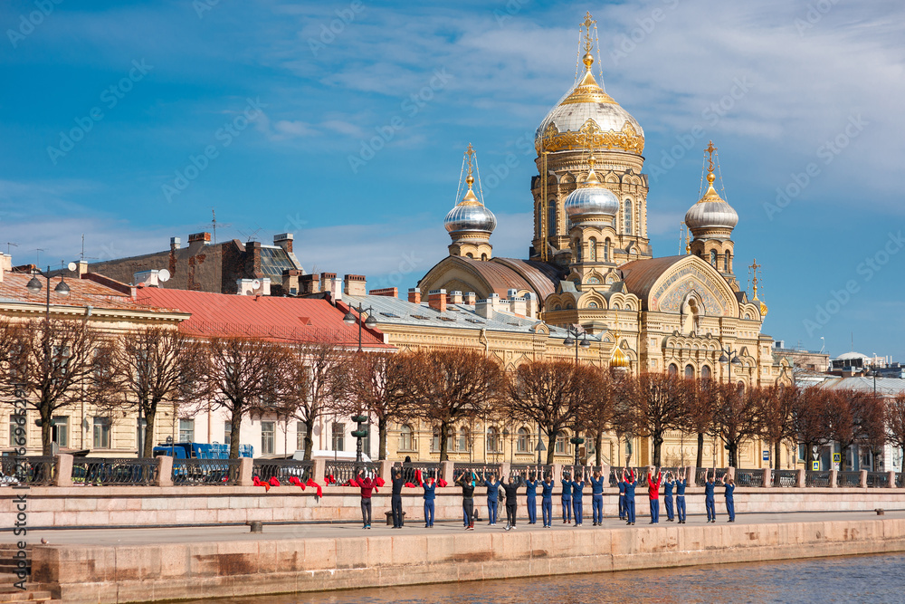 Fototapeta premium Group of people in uniform perform stretching exercises on the granite Neva embankment in Saint Petersburg, with the golden domed Assumption Church visible across the water