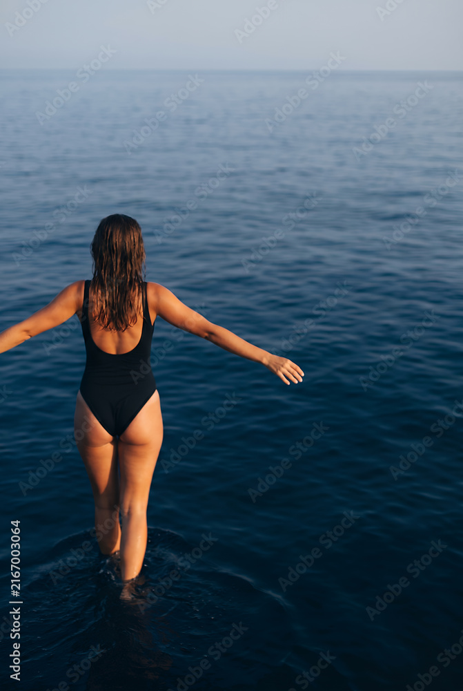Fototapeta premium Woman in black swimsuit wading in calm turquoise sea at golden sunset