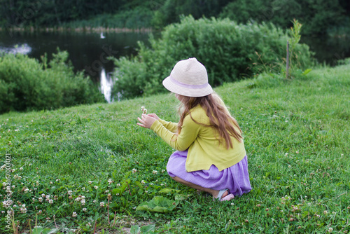 Canvastavla A young girl with long brown hair, wearing a yellow sweater and a pink hat, crouches on green grass, picking flowers near a calm lake surrounded by trees