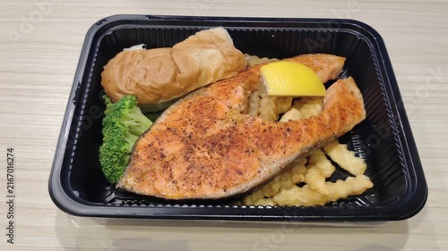 Grilled salmon bento meal served in a black takeaway container, featuring seasoned salmon fillet, tempura vegetables, broccoli, lemon wedge, French fries and a soft bread roll on a wooden table.