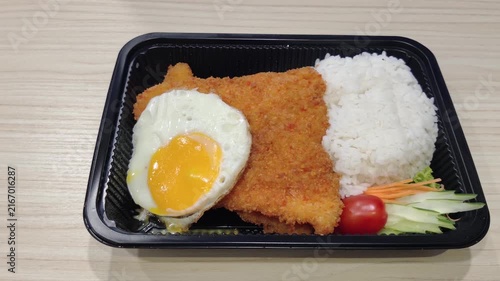 Breaded fried fish fillet served with steamed white rice, a sunny side up fried egg, cucumber, carrot, lettuce, and cherry tomato in a black takeaway food container.