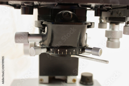 Detailed close-up of the mechanical and optical components of an old scientific microscope, featuring multiple objectives, a graduated stage mechanism and focusing controls.