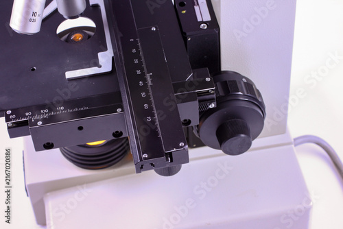 Close-up of a laboratory microscope stage with precision measurement scales, specimen holder, mechanical controls and illumination system for accurate sample positioning.