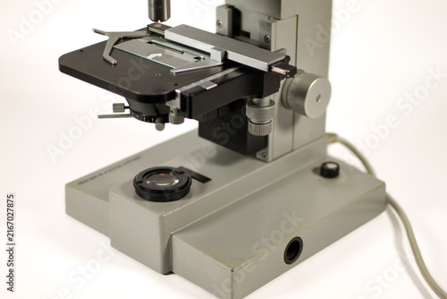 Detailed view of a classic microscope stage with specimen slide, metal clamps, precision positioning mechanism, focusing controls and sturdy illuminated base.