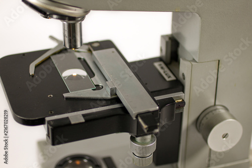 Close-up view of a vintage laboratory microscope stage with a metal slide holder, precision measurement scale, mechanical positioning controls and objective lens