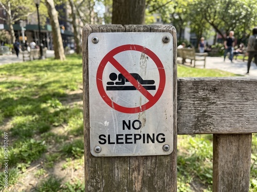No Sleeping Signage Mounted on a Wooden Post in a Public City Park with Blurred Background People and Greenery