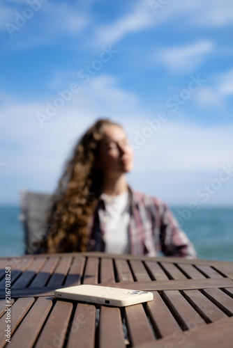 Foto Smartphone in the foreground with a woman relaxing by the sea in soft focus behind it