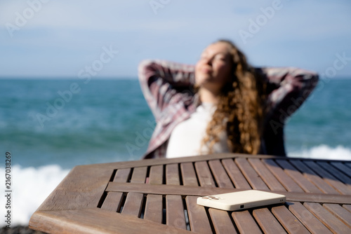 Cuadro en lienzo Smartphone in the foreground and a woman relaxing by the seaside in the background