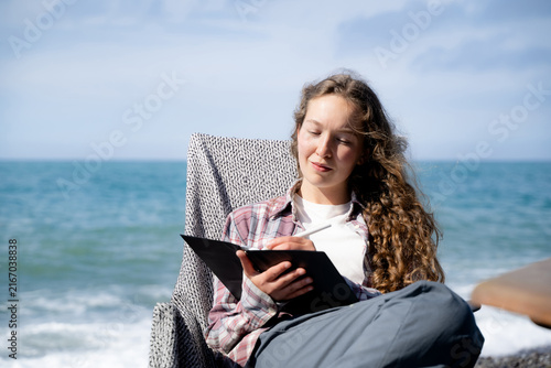 Принти на полотні Young woman keeping a gratitude journal by the sea on a sunny day, writing in a notebook while enjoying a quiet outdoor moment
