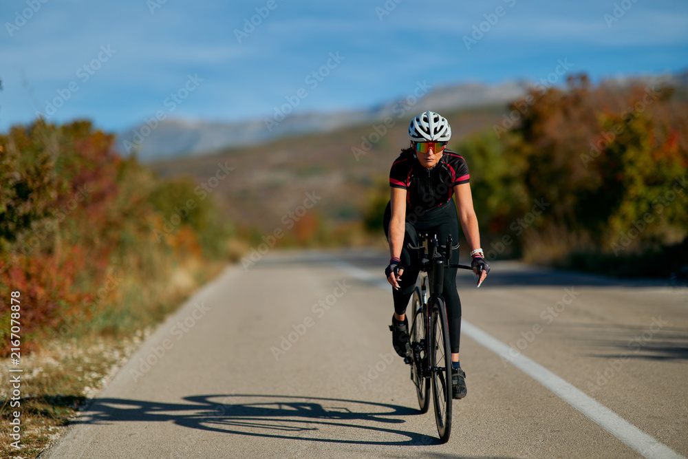 Naklejka premium Female Road Cyclist Training on Open Country Road in Autumn With Helmet and Sunglasses