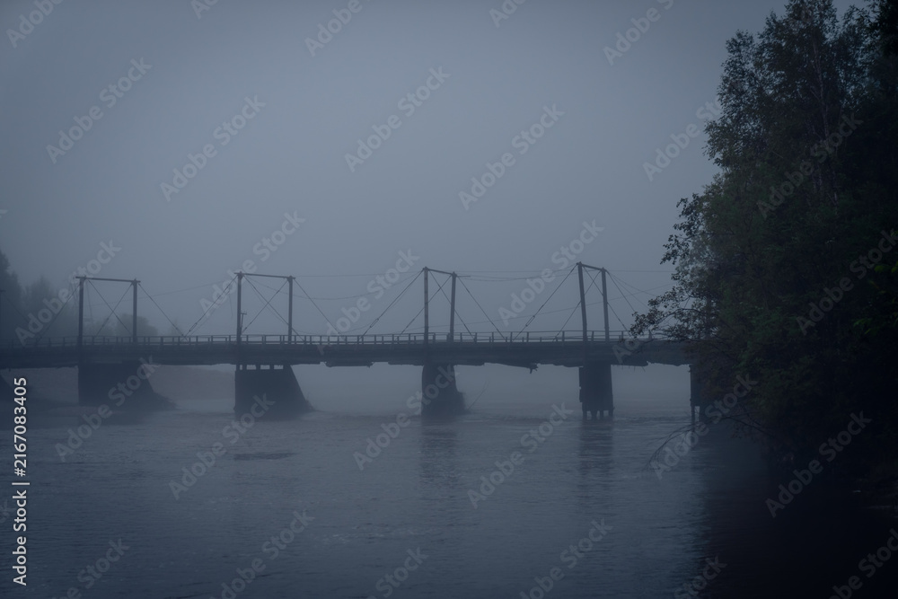 Fototapeta premium Foggy suspension bridge spanning calm river with muted tones and trees partially visible through dense mist