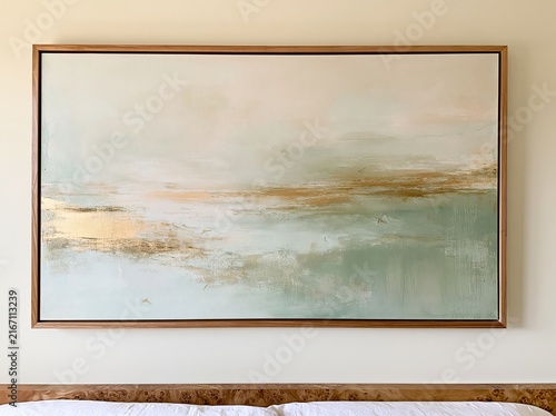 Large abstract landscape painting featuring soft blue and gold tones, framed in wood, displayed on a light-colored wall above a wooden bed