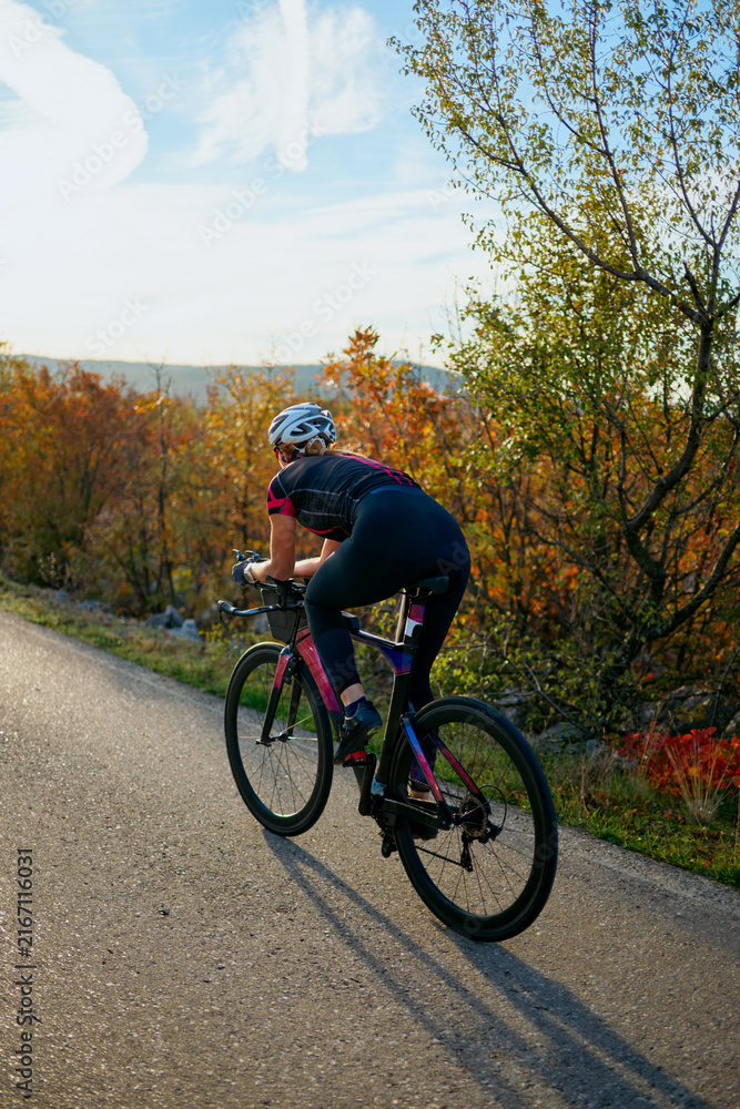 Naklejka premium Female Cyclist Training on Quiet Country Road at Sunset in Autumn Landscape With Scenic Trees