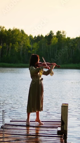 Young woman playing violin on a wooden lakeside pier at golden hour, creating peaceful music surrounded by calm water and lush green forest scenery