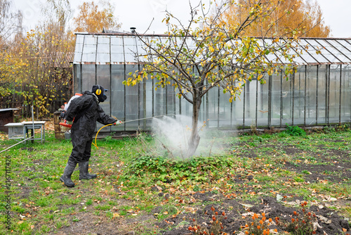 man in mask and overalls sprays garden with against pests in autumn.