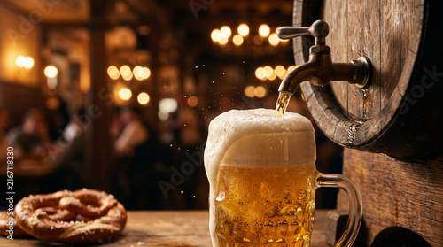 Pouring beer from wooden barrel tap into glass mug with pretzel in tavern